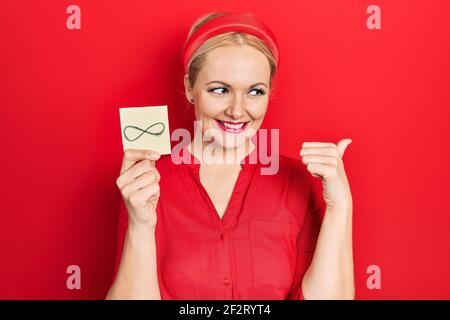 Jeune femme blonde portant un symbole de rappel infini pointant vers le haut sur le côté souriant heureux avec la bouche ouverte Banque D'Images