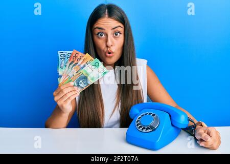 Belle femme hispanique avec téléphone vintage tenant des dollars australiens effrayée et stupéfaite avec la bouche ouverte pour la surprise, le visage incrédulé Banque D'Images