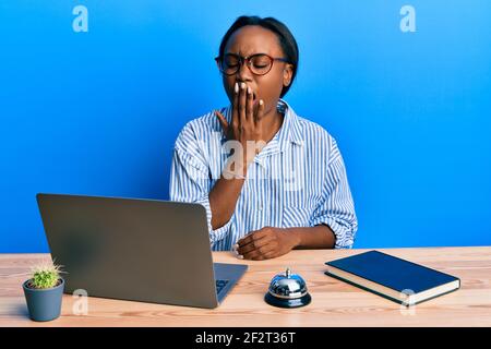 La jeune femme africaine travaillant à la réception de l'hôtel en utilisant un ordinateur portable s'ennuie bâillements fatigués couvrant la bouche avec la main. Agité et somnolence. Banque D'Images