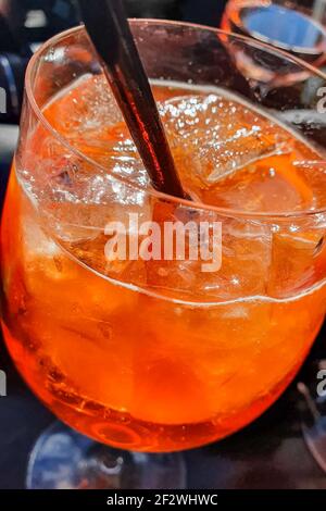 Verre d'Aperol Spritz avec une paille noire, servi sur une terrasse de bar donnant sur la ville de Manarola, Cinque Terre, Italie. Banque D'Images