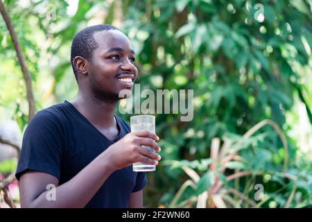 Homme africain tenant et buvant un verre d'eau dans fond vert naturel avec sourire et heureux Banque D'Images