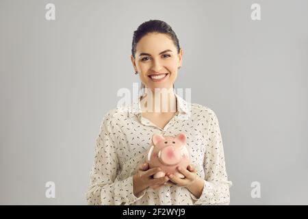 Une femme heureuse avec un sourire joyeux tient une banque de porc rose dans sa main debout sur un fond gris. Banque D'Images