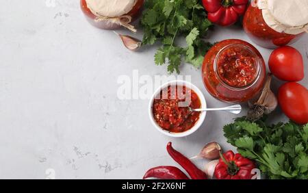 L'adjika arménienne de poivron rouge, de tomates, d'ail, de coriandre et de persil dans un bol et des bocaux sur fond gris. Vue de dessus. Espace pour le texte Banque D'Images