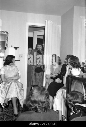 années 1950, historique, quelle surprise, une jeune femme dans un manteau élégant et tenant un petit sac à main, ouvre la porte à un appartement et à sa grande surprise, un groupe de ses amies attendent à l'intérieur! Banque D'Images