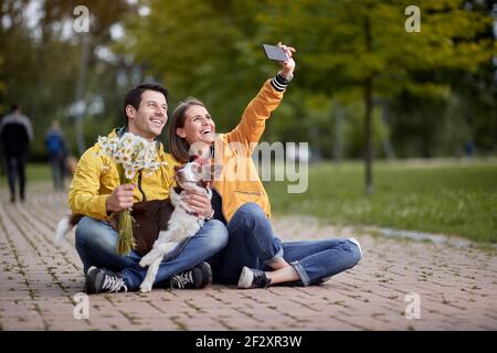 une jeune femme caucasienne prend le selfie avec son petit ami et son chien sur un tour, assis sur un chemin dans le parc Banque D'Images