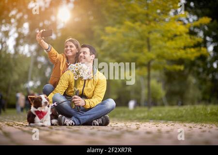 couple caucasien avec un chien assis sur un chemin dans le parc avec les jambes croisées, prenant selfie avec leur chien Banque D'Images