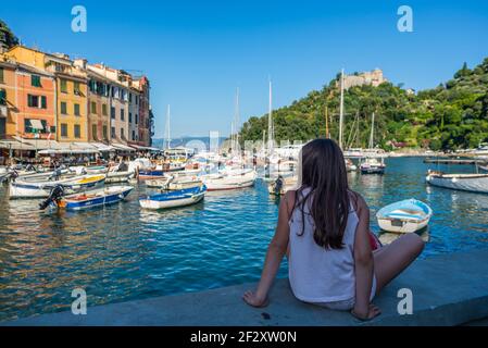 Une jeune fille aux cheveux longs bruns est assise sur un mur et regarde la baie de Portofino. Le château Brown en arrière-plan au sommet d'une colline Banque D'Images