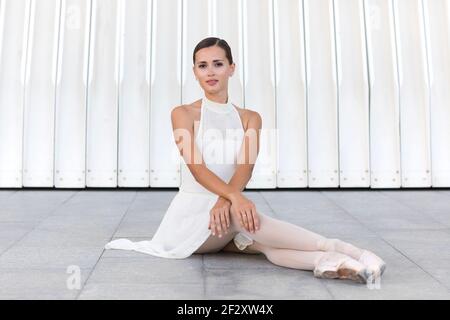Jeune danseuse de ballet charmante en robe blanche et pointe chaussures regardant l'appareil photo en étant assis dans la rue Banque D'Images