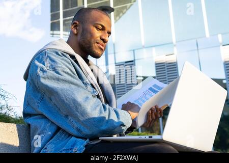 De l'employé à distance ethnique mâle d'un niveau inférieur à l'adulte avec document et netbook installé en ville Banque D'Images