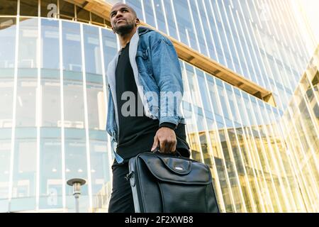 Vue d'en dessous de côté de l'homme ethnique adulte dans une ambiance décontractée tenue avec porte-documents en cuir se promenant en ville tout en regardant loin Banque D'Images