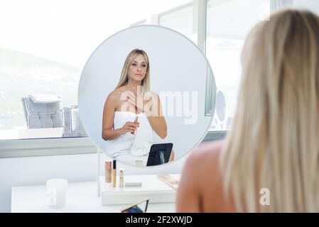 Charmante femme assise devant le miroir et appliquant de la crème à la maison Banque D'Images