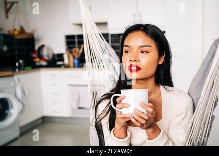 Jeune femme hispanique pensive assise dans un hamac dans une cuisine moderne avec une boisson chaude pendant la journée Banque D'Images