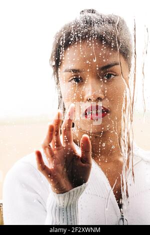 À travers le verre de l'ethnie pensive femelle avec des lèvres vives regardant à l'appareil photo tout en touchant les drops Banque D'Images