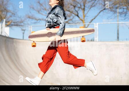 Vue latérale d'une petite jeune femme ravie et élégante vêtements sautant sur le parc de skate en béton Banque D'Images