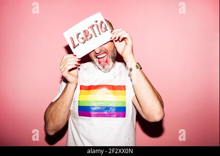 Croiser l'homme homosexuel barbu rebelle avec des lèvres rouges et la manucure faire de la grimace tout en montrant et en couvrant la face avec du papier Texte LGBTIQ contre p Banque D'Images