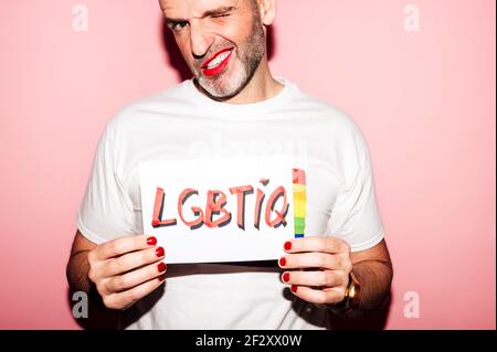Croiser l'homme homosexuel barbu rebelle avec des lèvres rouges et la manucure Faire de la grimace tout en montrant du papier avec du texte LGBTIQ contre du rose arrière-plan Banque D'Images