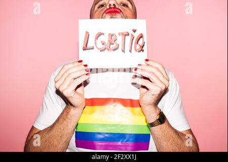 Homme homosexuel barbu rebelle aux lèvres rouges et manucure Grimace tout en montrant et en couvrant la face avec du papier avec LGBTIQ texte contre rose b Banque D'Images