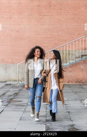 Des amies ravies avec des sourires crasseux portant des vêtements tendance loin tout en marchant ensemble en ville Banque D'Images
