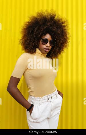 Jeune femme ethnique avec une coiffure afro et des lunettes de soleil debout mur jaune Banque D'Images