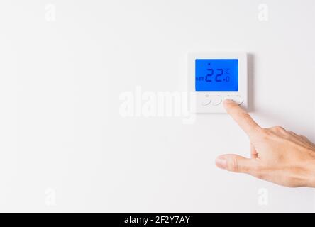 Main d'homme caucasien réglant la température d'un thermostat blanc à vingt-deux degrés avec l'affichage bleu rétroéclairé sur un mur blanc Banque D'Images
