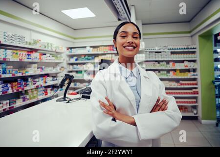 Fière jeune femme avec les mains repliées dans un manteau debout à l'intérieur pharmacie près du comptoir Banque D'Images