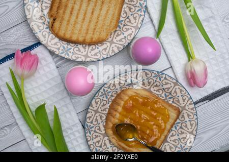 Œuf de Pâques de couleur rose dans l'assiette sur une table de petit déjeuner avec confiture et tulipes blanches roses et pain grillé. Banque D'Images