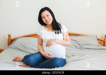 Femme enceinte chinoise asiatique souriante assise sur un lit touchant le ventre. Je m'attends à une jeune femme en T-short blanc et jeans bleus à la maison. Santé heureux Banque D'Images