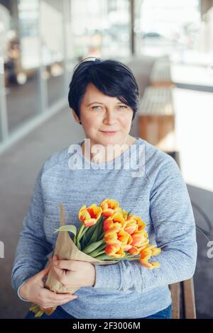 Adulte d'âge moyen mature Femme caucasienne avec de courts cheveux noirs assis sur le banc à l'extérieur et tenant des fleurs de tulipe rouge jaune. Printemps et printemps Banque D'Images