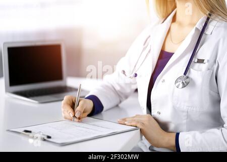 Une femme-médecin inconnue prescrit des médicaments à son patient, à l'aide d'un presse-papiers, tout en étant assise au bureau dans son cabinet ensoleillé. Femme Banque D'Images