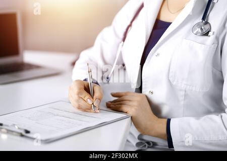 Une femme-médecin inconnue prescrit des médicaments à son patient, à l'aide d'un presse-papiers, tout en étant assise au bureau dans son cabinet ensoleillé. Femme Banque D'Images