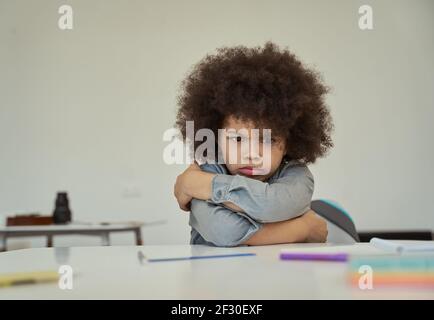 Un petit écolier mécontent aux cheveux d'afro qui ont l'air plein de ressentiment, se froissant en étant assis avec les bras croisés à la table dans la salle de classe de l'école primaire Banque D'Images