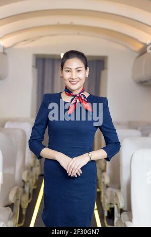 Un agent de bord asiatique se posant avec le sourire au milieu de la allée à l'intérieur du siège passager de l'avion sur l'arrière-plan Banque D'Images