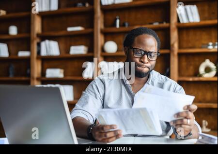 Confondu un jeune étudiant afro-américain ou un entrepreneur intelligent en lunettes vérifiant les numéros des journaux, assis à la table dans une bibliothèque et tenant des documents, relisant l'accord avec le client Banque D'Images