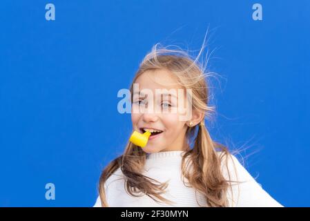 Studio portrait d'une jeune fille heureuse et souriante célébrant la fête d'anniversaire, tenant le souffleur de fête dans la bouche s'amusant, isolé sur le bleu. Photo de haute qualité Banque D'Images