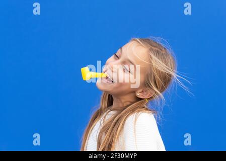 Studio portrait d'une jeune fille heureuse et souriante célébrant la fête d'anniversaire, tenant le souffleur de fête dans la bouche s'amusant, isolé sur le bleu. Photo de haute qualité Banque D'Images