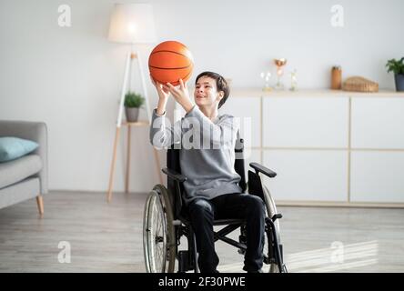 Joyeux adolescent handicapé jouant avec le basket-ball, assis en fauteuil roulant à la maison Banque D'Images