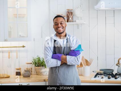 Portrait d'un jeune homme noir en tablier et gants en caoutchouc sourire à l'appareil photo dans la cuisine Banque D'Images