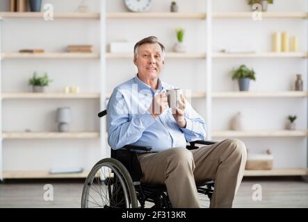 Homme handicapé âgé positif en fauteuil roulant buvant du café ou du thé à la maison Banque D'Images