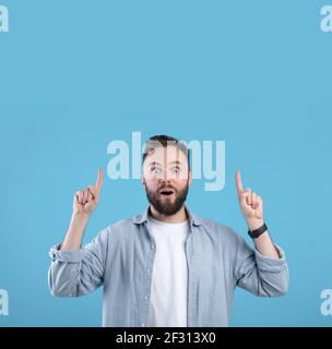 Beau barbu homme ayant choqué l'expression du visage, pointant sur un espace vide au-dessus de sa tête sur fond bleu studio Banque D'Images