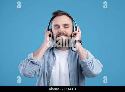 Beau barbu dans une tenue décontractée à l'écoute de la musique, portant des écouteurs sur fond bleu studio Banque D'Images