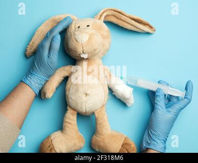 Lapin en peluche avec une patte bandée et deux mains femelles d'un médecin injectant une injection sur fond bleu Banque D'Images