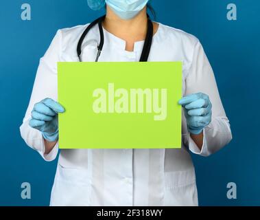 Femme médecin dans un manteau médical blanc, un masque jetable et un capuchon se tient sur un fond bleu et tient une feuille vierge verte de p Banque D'Images