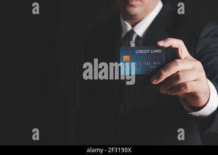 homme d'affaires en costume noir et cravate grise montrant une carte de crédit sur fond noir. Concept de banque de paiement Banque D'Images