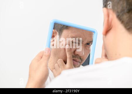 Un homme d'âge moyen regardant dans le miroir et inquiet des imperfections et des rides sur son visage sur fond blanc, concept de soins de santé Banque D'Images