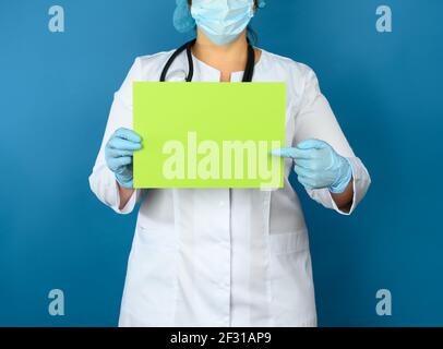 Femme médecin dans un manteau médical blanc, un masque jetable et un capuchon se tient sur un fond bleu et tient une feuille vierge verte de p Banque D'Images