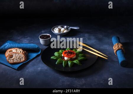 Variété de salade Caprese, tranches de tomates mûres fraîches, mini-mozzarella, feuilles de basilic frais et caviar balsamique. Tableau d'arrière-plan sombre. Banque D'Images