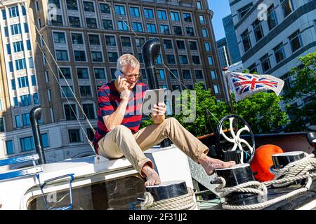 Bureau à domicile homme d'affaires mature à l'extérieur de la maison bateau bureau parler sur smartphone regardant l'ordinateur tablette bureaux de Canary Wharf derrière Londres E14 Banque D'Images