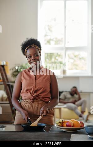 Jeune femme souriante d'origine africaine qui vous regarde pendant debout près d'une cuisinière électrique dans la cuisine et de faire frire quelque chose pour sa famille Banque D'Images