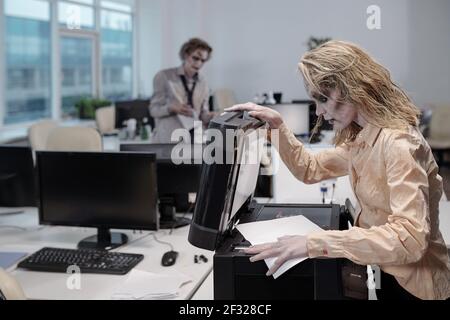 Jeune femme avec zombie Greasepaint sur le visage et les mains debout par la machine xerox et l'ouverture du couvercle avant de faire des copies de documents Banque D'Images