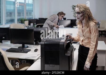 Jeune femme d'affaires zombie debout par la machine xerox tout en faisant des copies de documents contre des hommes morts collègue avec des papiers Banque D'Images